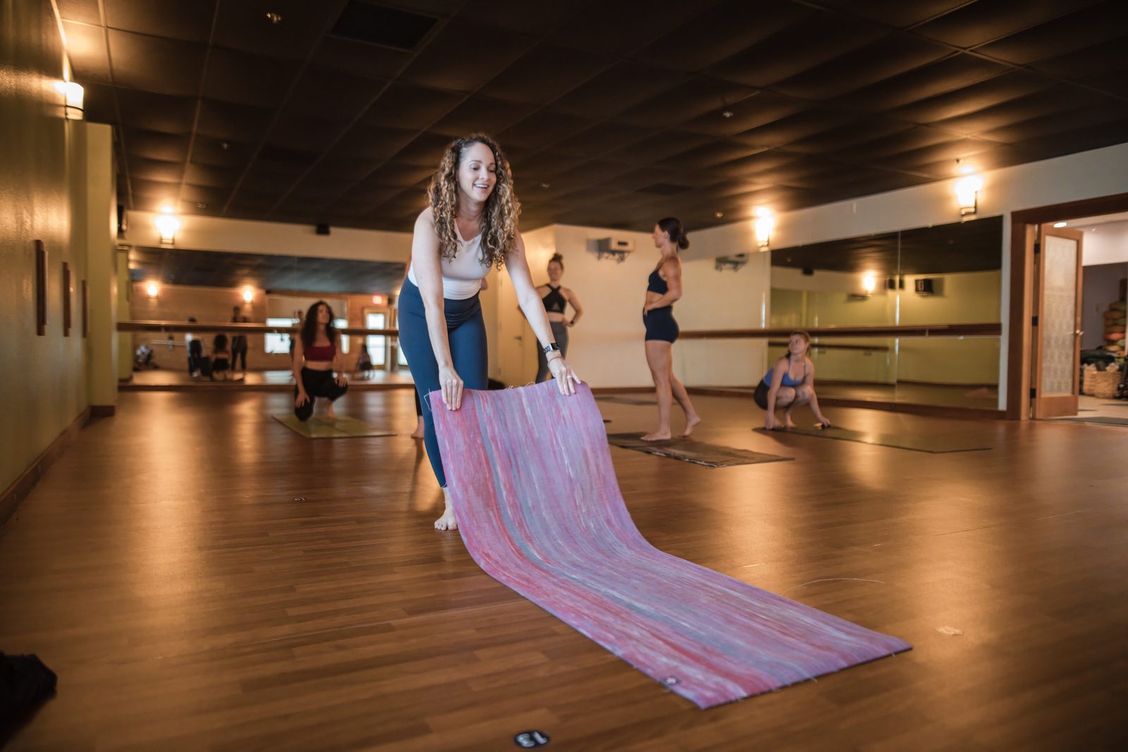 An instructor smiles as she rolls out her mat, while students set up behind her